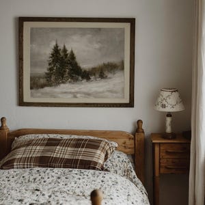 Puede incluir: Un cuadro de paisaje enmarcado de pinos en un campo nevado cuelga sobre una estructura de cama de madera. La cama cuenta con una almohada de cuadros marrones y un edred&oacute;n floral blanco. Junto a la cama hay una mesita de noche con una l&aacute;mpara.