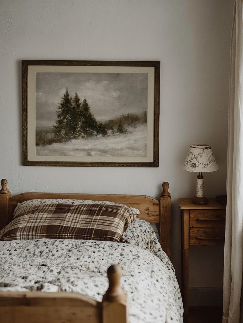 Puede incluir: Un cuadro de paisaje enmarcado de pinos en un campo nevado cuelga sobre una estructura de cama de madera. La cama cuenta con una almohada de cuadros marrones y un edred&oacute;n floral blanco. Junto a la cama hay una mesita de noche con una l&aacute;mpara.