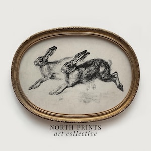 Puede incluir: Una impresión enmarcada ovalada que muestra dos liebres corriendo, en un estilo vintage. La obra de arte es en blanco y negro, con las liebres representadas en pleno salto. El marco es dorado y la impresión tiene el texto "NORTH PRINTS art collective" en la parte inferior.