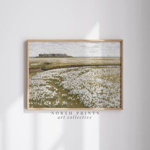 May include: A framed print of a landscape painting depicting a field of white flowers with a distant view of a small town. The painting is in a light brown frame and has a white background.