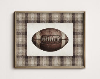 May include: A framed print featuring a vintage-style football. The football is brown with white stripes and laces, set against a cream background. The frame has a gold finish and a plaid fabric border, creating a classic, rustic aesthetic.
