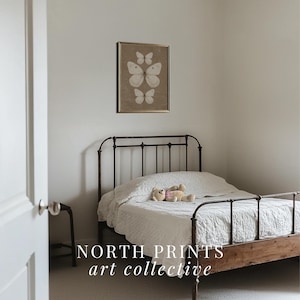 May include: A white metal bed frame with a white linen headboard and a white linen comforter. A stuffed animal is lying on the bed. A framed print of butterflies on a brown background hangs on the wall above the bed. The text "NORTH PRINTS art collective" is printed below the bed.