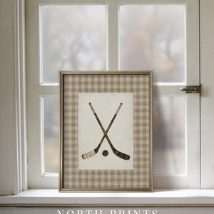 May include: Framed art print featuring crossed hockey sticks and a puck. The artwork is set against a beige and white checkered background, framed in gold. The text "NORTH PRINTS art collective" is at the bottom.