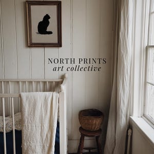 Peut inclure: Une &oelig;uvre encadr&eacute;e repr&eacute;sentant une silhouette de chat noir est accroch&eacute;e au-dessus d'un berceau blanc avec une couverture cr&egrave;me. Un petit tabouret en bois supporte un panier en osier. Le texte "NORTH PRINTS art collective" est visible.