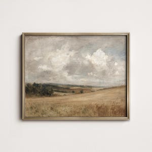 May include: A framed landscape painting depicts a field of golden wheat under a cloudy sky. The artwork features a muted color palette with shades of brown, beige, and gray. The painting is enclosed in a gold-colored frame.