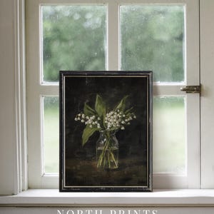 May include: A framed painting of a bouquet of white lily of the valley flowers in a clear glass vase on a dark brown background. The painting is in a black frame and is sitting on a white windowsill.