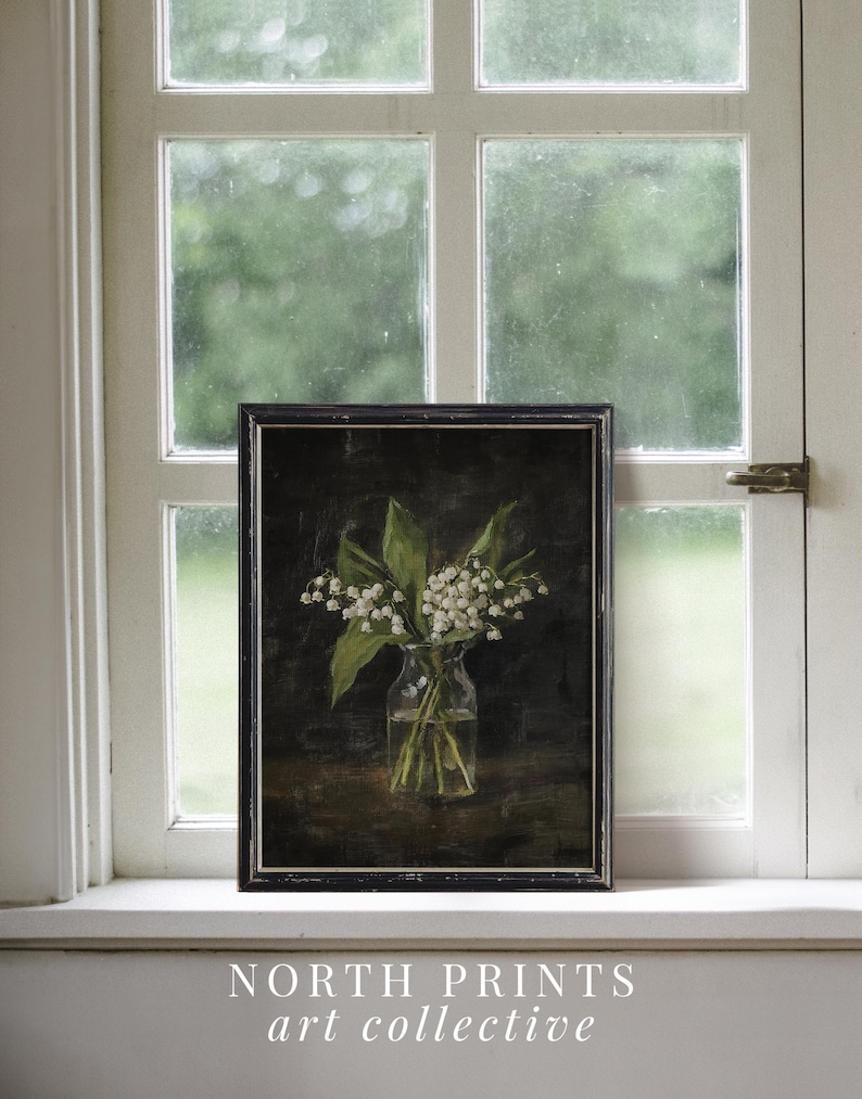 May include: A framed painting of a bouquet of white lily of the valley flowers in a clear glass vase on a dark brown background. The painting is in a black frame and is sitting on a white windowsill.