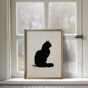 Peut inclure: Une affiche encadr&eacute;e repr&eacute;sentant une silhouette de chat noir. Le chat est assis, la queue enroul&eacute;e. L'affiche est dans un cadre dor&eacute;, expos&eacute;e sur un rebord de fen&ecirc;tre. Le texte "NORTH PRINTS art collective" est en bas.