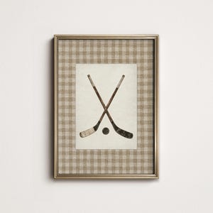 May include: Framed print featuring two crossed hockey sticks and a puck. The artwork is set against a cream-colored background with a gold-colored frame. The frame has a tan and white checkered pattern.