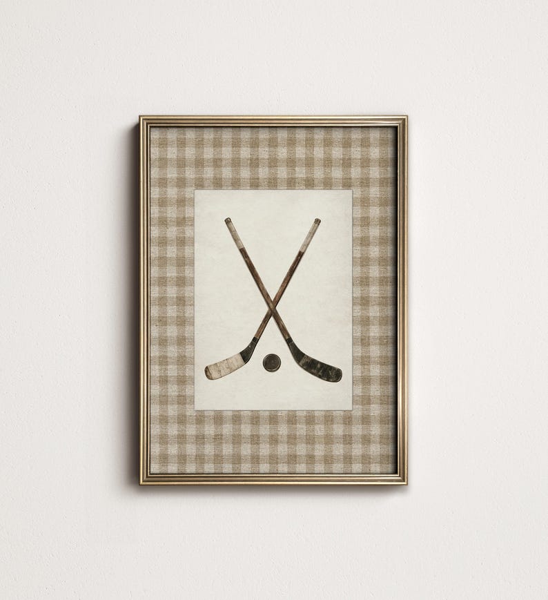 May include: Framed print featuring two crossed hockey sticks and a puck. The artwork is set against a cream-colored background with a gold-colored frame. The frame has a tan and white checkered pattern.
