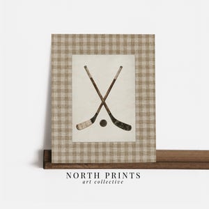 May include: A framed print featuring two crossed hockey sticks and a puck. The artwork is set against a cream-colored background and framed with a tan and white checkered pattern. The text "NORTH PRINTS art collective" is at the bottom.