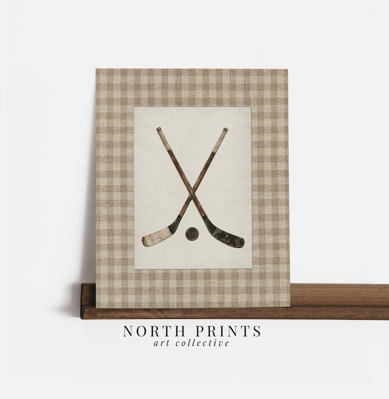 May include: A framed print featuring two crossed hockey sticks and a puck. The artwork is set against a cream-colored background and framed with a tan and white checkered pattern. The text "NORTH PRINTS art collective" is at the bottom.
