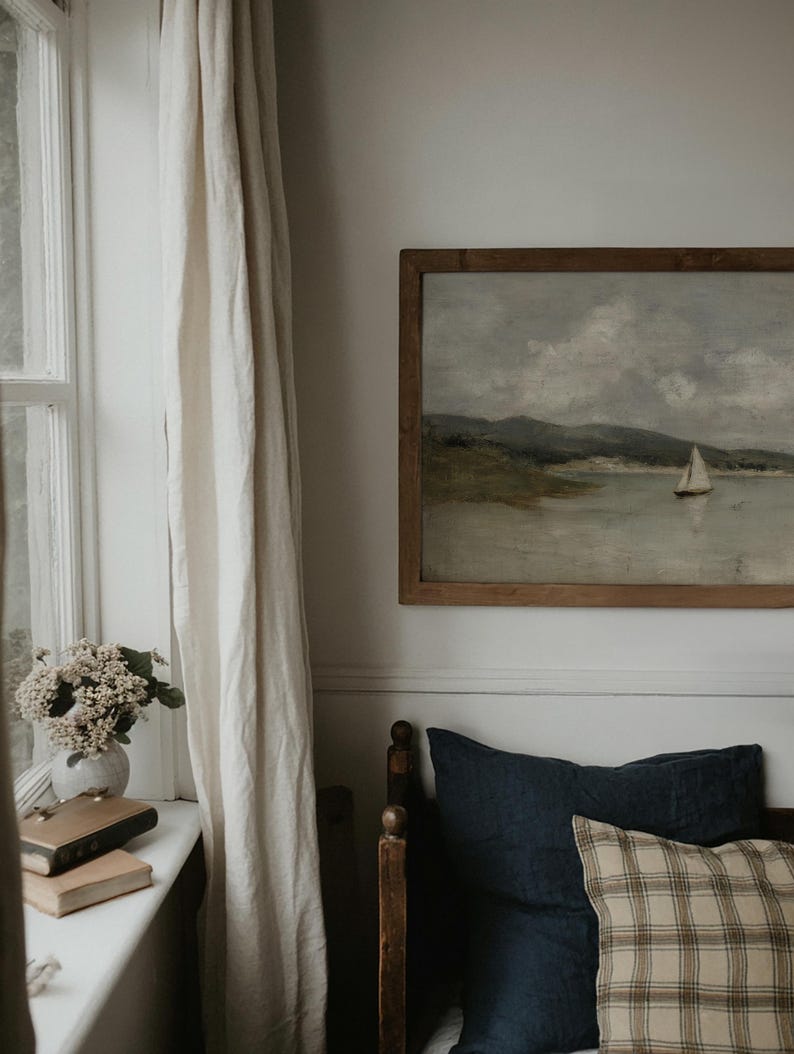 K&ouml;nnte beinhalten: Ein gerahmtes Landschaftsgem&auml;lde eines Segelbootes auf dem Wasser h&auml;ngt &uuml;ber einem Bett mit marineblauen und karierten Kissen. Links vom Bett befindet sich ein Fenster mit wei&szlig;en Vorh&auml;ngen und eine Vase mit Blumen.