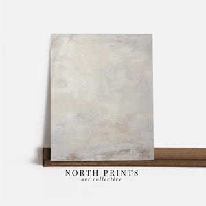 May include: Abstract painting with a white, grey, and beige colour scheme. The painting has a textured surface and a subtle brushstroke pattern. The painting is framed and hanging on a wooden shelf. The text "NORTH PRINTS art collective" is visible below the painting.