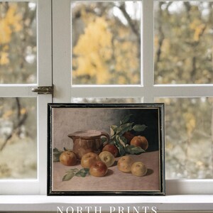 May include: A framed still life painting of a brown pitcher and apples on a table. The painting is set against a blurred background of fall foliage.