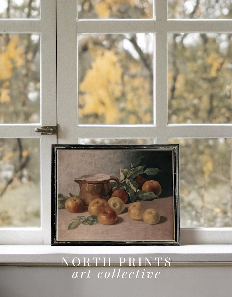May include: A framed still life painting of a brown pitcher and apples on a table. The painting is set against a blurred background of fall foliage.