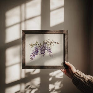 May include: A framed watercolor print featuring a sprig of purple wisteria blossoms. The artwork is set in a dark brown wooden frame and held against a neutral wall, with sunlight casting shadows.
