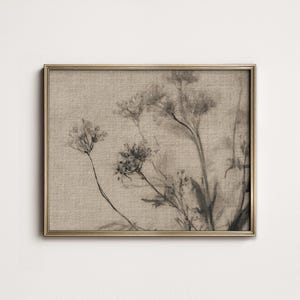 Puede incluir: Una impresión botánica enmarcada con un arreglo floral en tonos sepia. La obra de arte presenta flores delicadas y tenues sobre un fondo beige texturizado. La impresión está encerrada en un marco delgado de color dorado, añadiendo un toque de elegancia.