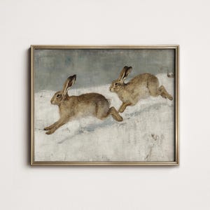 May include: A framed vintage print depicts two brown hares running across a snowy landscape. The artwork has a muted color palette with a gold-colored frame, evoking a classic, rustic aesthetic. The hares are the focal point.