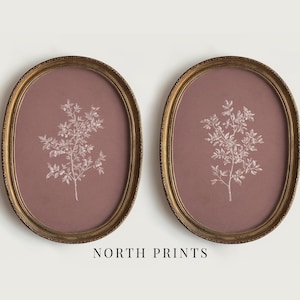 Peut inclure: Deux impressions botaniques encadrées. Chaque cadre ovale a une finition dorée et un fond mauve. Un motif floral blanc et délicat est centré sur chaque impression. Le texte "NORTH PRINTS" est au bas de l'image.