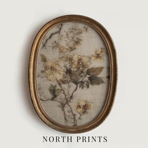 Op de afbeelding: Een ovaalvormige ingelijste print met een botanisch bloemenontwerp. Het kunstwerk toont een gedempt kleurenpalet met beige, gele en groene tinten. De lijst is goudkleurig met ingewikkelde details. De tekst "NORTH PRINTS" staat onderaan.