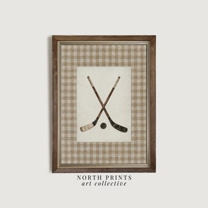May include: Framed print featuring crossed hockey sticks and a puck. The artwork is set against a cream-colored background with a brown and tan plaid pattern. The frame is a dark brown color. The text "NORTH PRINTS art collective" is at the bottom.