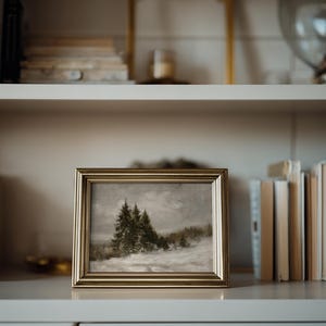 Puede incluir: Un cuadro de paisaje rectangular en un marco de metal dorado, que representa una escena nevada con &aacute;rboles de hoja perenne oscuros bajo un cielo nublado y apagado. La obra presenta una textura suave con una paleta de tonos blancos, grises y verde oscuro.