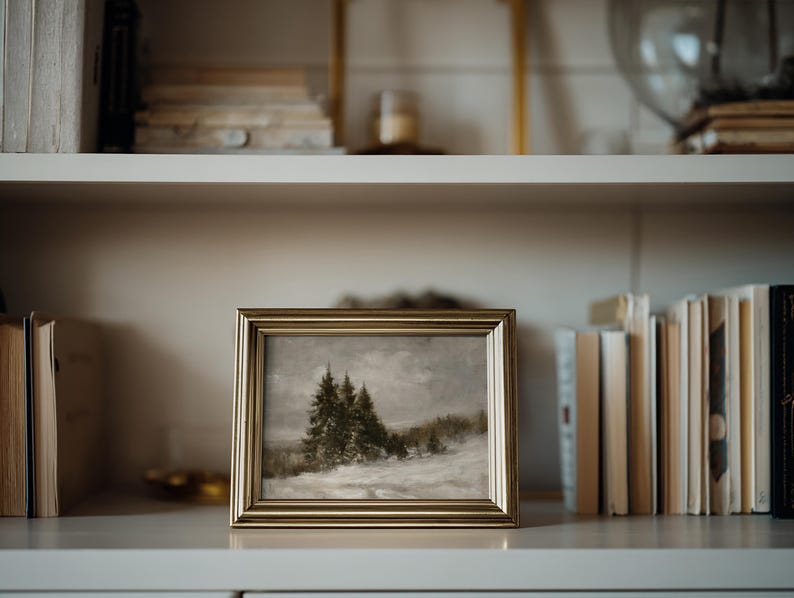 Puede incluir: Un cuadro de paisaje rectangular en un marco de metal dorado, que representa una escena nevada con &aacute;rboles de hoja perenne oscuros bajo un cielo nublado y apagado. La obra presenta una textura suave con una paleta de tonos blancos, grises y verde oscuro.