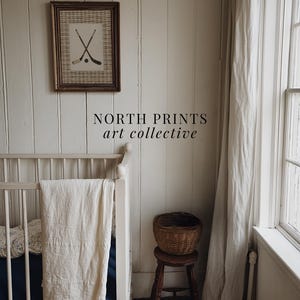 May include: A framed print featuring crossed hockey sticks and a puck hangs on a white paneled wall. The text "NORTH PRINTS art collective" is below the print. A white crib with a blanket and a small wooden stool with a basket are in the foreground.