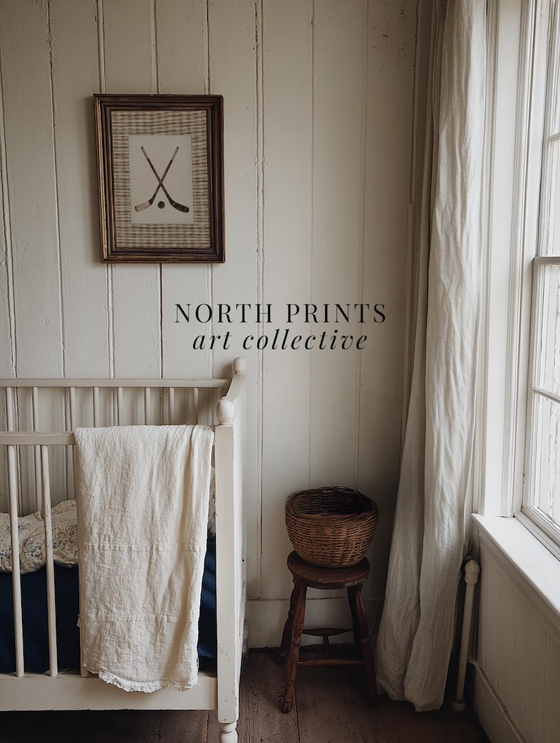 May include: A framed print featuring crossed hockey sticks and a puck hangs on a white paneled wall. The text "NORTH PRINTS art collective" is below the print. A white crib with a blanket and a small wooden stool with a basket are in the foreground.