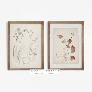 May include: Two framed botanical prints, one with a black and white drawing of a flower and the other with a watercolor painting of leaves in shades of pink and brown. The frames are wood with a light brown finish. The prints are titled "NORTH PRINTS vintage art collective".