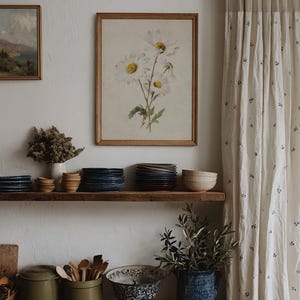 Op de afbeelding: Een rustieke keuken met een houten plank met stapels borden, kommen en decoratieve items. Een ingelijste bloemenprint hangt aan de muur, aangevuld met een gordijn met een subtiel blauw bloemmotief. De sfeer is warm en uitnodigend.