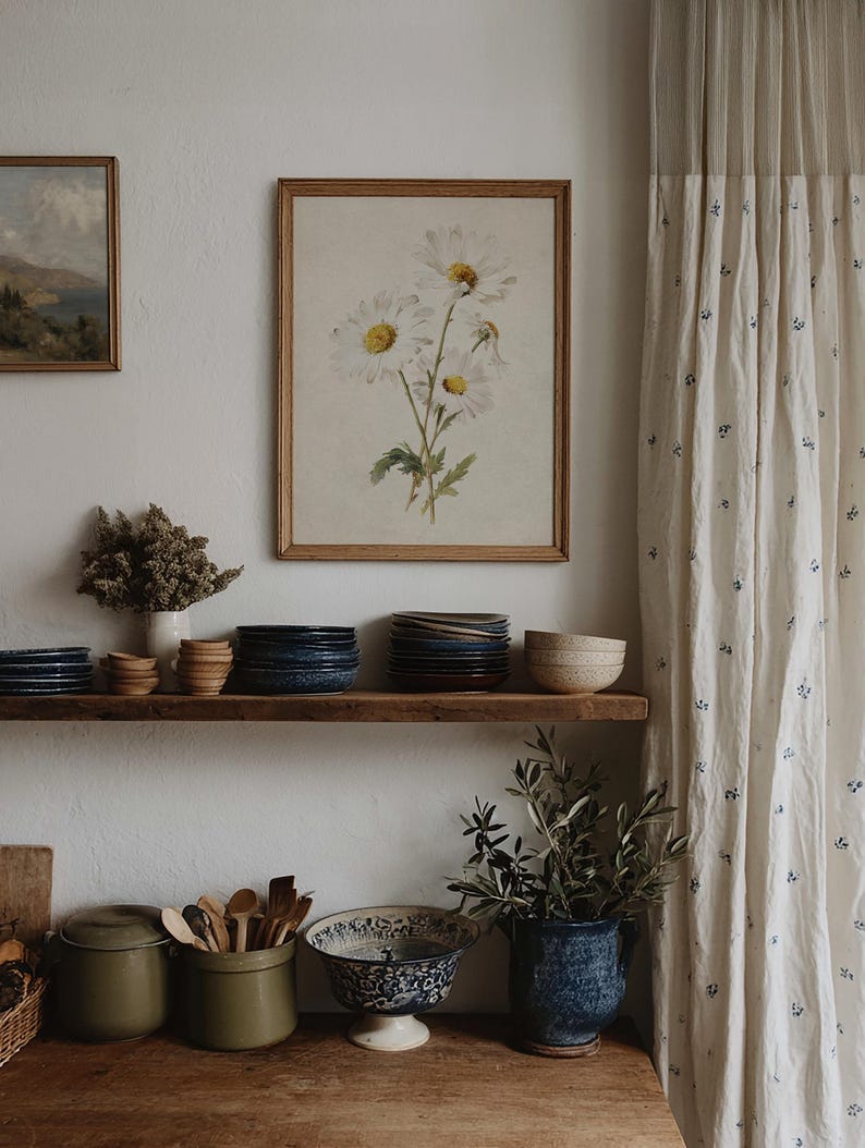 Op de afbeelding: Een rustieke keuken met een houten plank met stapels borden, kommen en decoratieve items. Een ingelijste bloemenprint hangt aan de muur, aangevuld met een gordijn met een subtiel blauw bloemmotief. De sfeer is warm en uitnodigend.