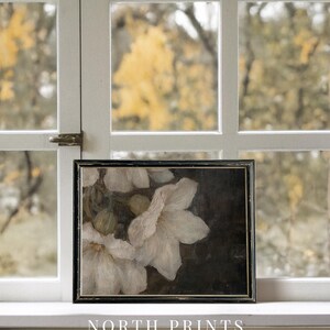 May include: A framed painting of white flowers on a dark background. The flowers are in focus, while the background is blurred. The painting is in a black frame and is sitting on a white windowsill.