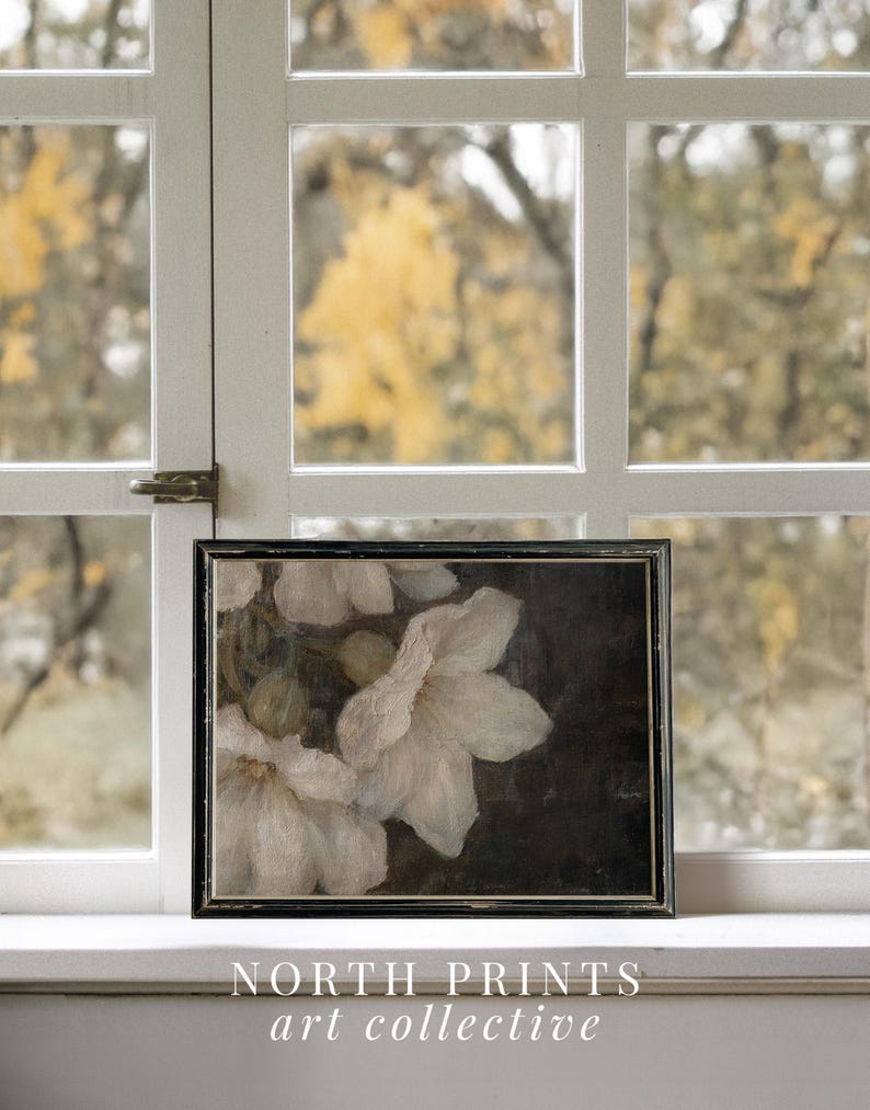 May include: A framed painting of white flowers on a dark background. The flowers are in focus, while the background is blurred. The painting is in a black frame and is sitting on a white windowsill.