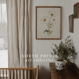 Op de afbeelding: Een ingelijste bloemenprint met witte madeliefjes en gele centra hangt aan een witte muur. Een houten wieg en een vintage houten dressoir met een potplant staan op de voorgrond. De tekst "NORTH PRINTS art collective" is zichtbaar.