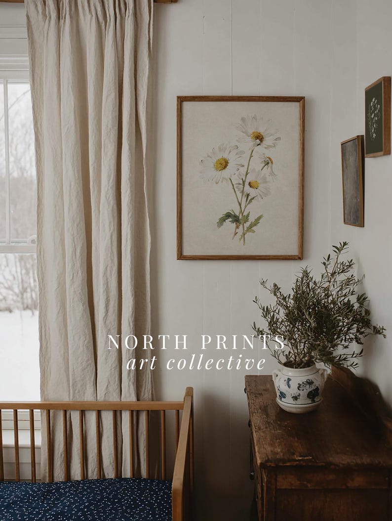 Op de afbeelding: Een ingelijste bloemenprint met witte madeliefjes en gele centra hangt aan een witte muur. Een houten wieg en een vintage houten dressoir met een potplant staan op de voorgrond. De tekst "NORTH PRINTS art collective" is zichtbaar.