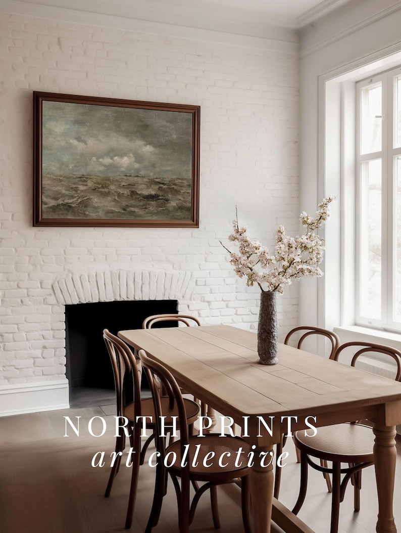 May include: A light wood dining table with six chairs, a vase of white flowers, and a framed painting of a seascape hanging on a white brick wall. The text "NORTH PRINTS art collective" is visible on the table.