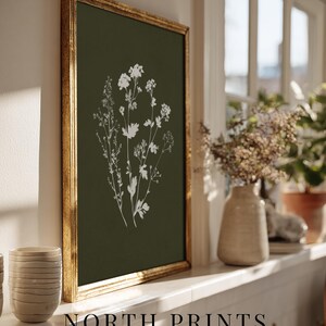 May include: A framed botanical print featuring white floral illustrations on a dark green background. The gold frame surrounds the artwork, which is displayed on a white windowsill. The text "NORTH PRINTS art collective" is at the bottom.