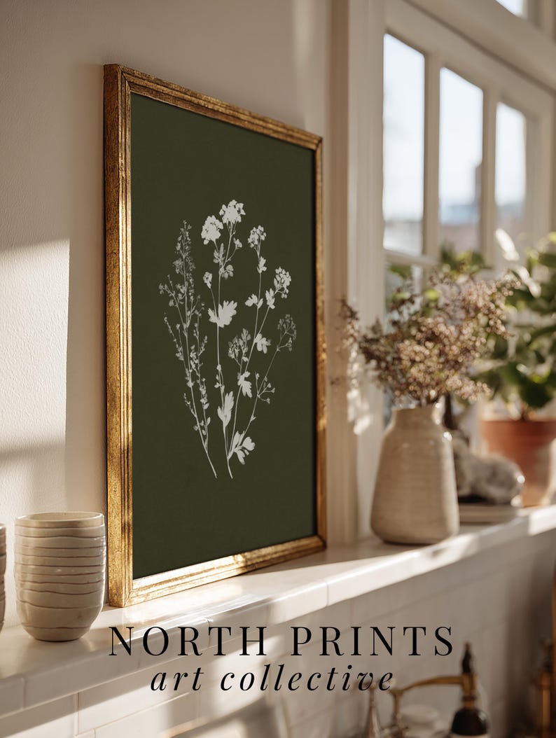 May include: A framed botanical print featuring white floral illustrations on a dark green background. The gold frame surrounds the artwork, which is displayed on a white windowsill. The text "NORTH PRINTS art collective" is at the bottom.