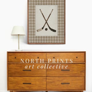 May include: A framed print featuring crossed hockey sticks and a puck. The artwork is set against a neutral background with a brown and beige plaid border. The print is displayed above a mid-century modern wooden dresser.