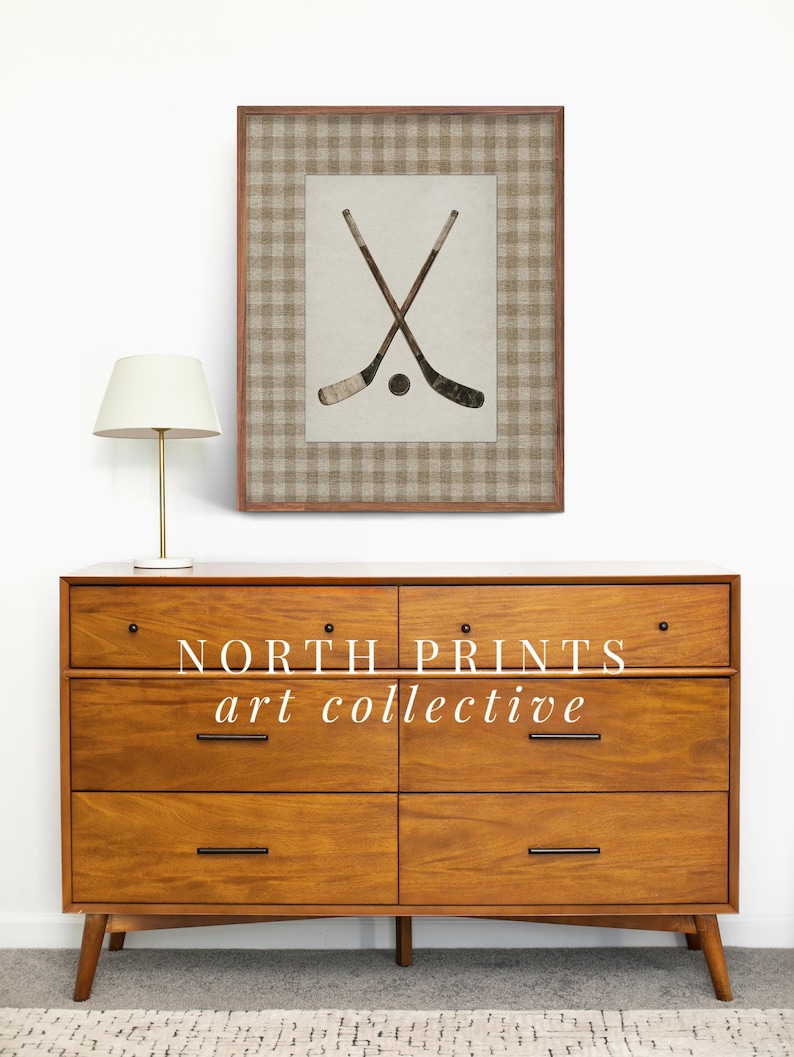 May include: A framed print featuring crossed hockey sticks and a puck. The artwork is set against a neutral background with a brown and beige plaid border. The print is displayed above a mid-century modern wooden dresser.