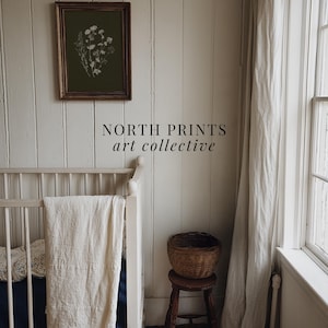 May include: An olive green framed print with a white floral design hangs on a white paneled wall. A white crib with a cream-colored blanket is in the foreground. A small wooden stool holds a woven basket. The text "NORTH PRINTS art collective" is below the print.