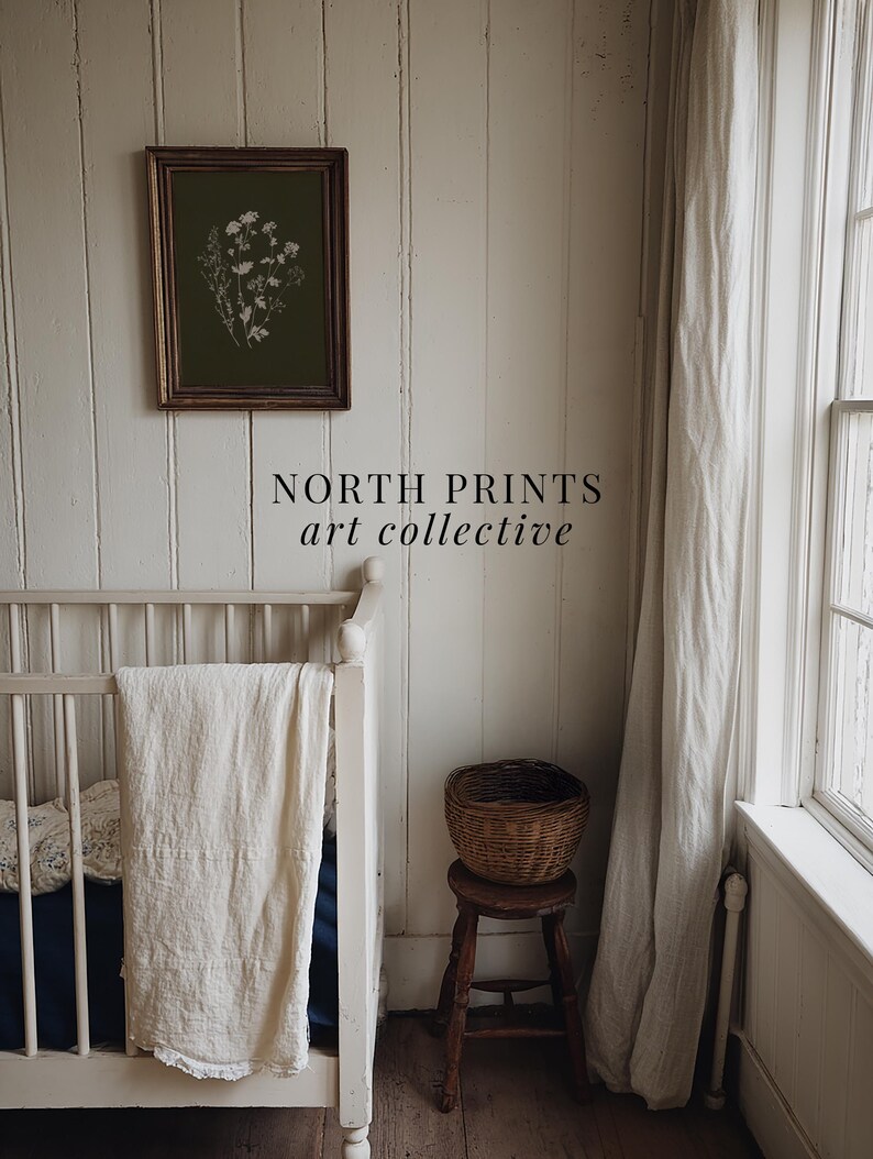 May include: An olive green framed print with a white floral design hangs on a white paneled wall. A white crib with a cream-colored blanket is in the foreground. A small wooden stool holds a woven basket. The text "NORTH PRINTS art collective" is below the print.