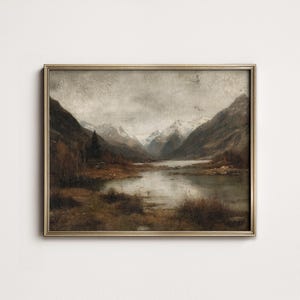 May include: A framed landscape painting of a mountain lake scene. The artwork uses a muted palette of browns, greys, and whites, with snow-covered mountains in the background. The painting is enclosed in a gold-coloured frame.