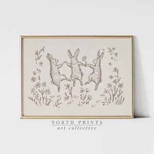 May include: A black and white line drawing of three rabbits dancing in a field of flowers. The image is framed in a light wood frame. The text "NORTH PRINTS art collective" is printed below the image.