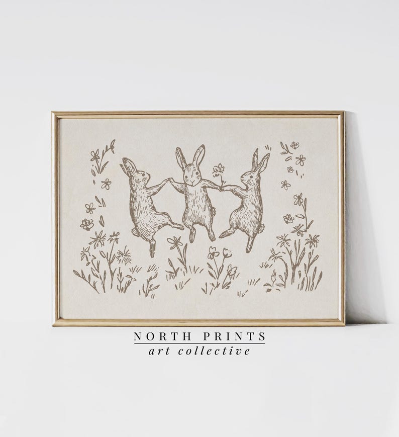 May include: A black and white line drawing of three rabbits dancing in a field of flowers. The image is framed in a light wood frame. The text "NORTH PRINTS art collective" is printed below the image.