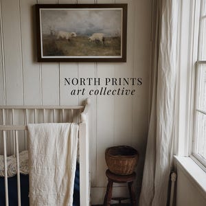 K&ouml;nnte beinhalten: Gerahmtes Landschaftsgem&auml;lde mit zwei Schafen auf einem Feld &uuml;ber einem wei&szlig;en Kinderbett. Ein kleiner Holzschemel tr&auml;gt einen Weidenkorb. Der Text "NORTH PRINTS art collective" ist darunter.
