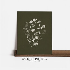 May include: A botanical print featuring a cluster of white wildflowers on a dark green background. The print is displayed on a wooden shelf. The text "NORTH PRINTS art collective" is printed at the bottom.