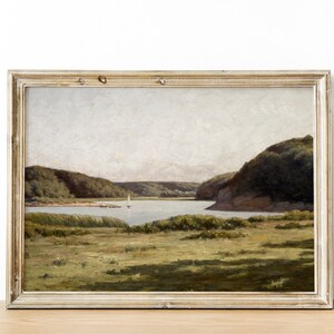 May include: A framed painting depicting a landscape scene with a lake, green hills, and a sailboat in the distance. The painting is set against a white wall.